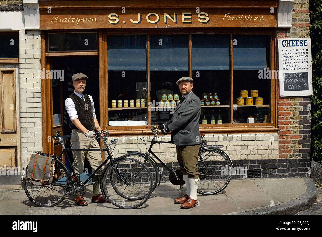 Beautiful gay couple at sunny day riding bike.Gay male couple standing beside retro bicycle in from of old bakery in Shoreditch, East London , England Stock Photo