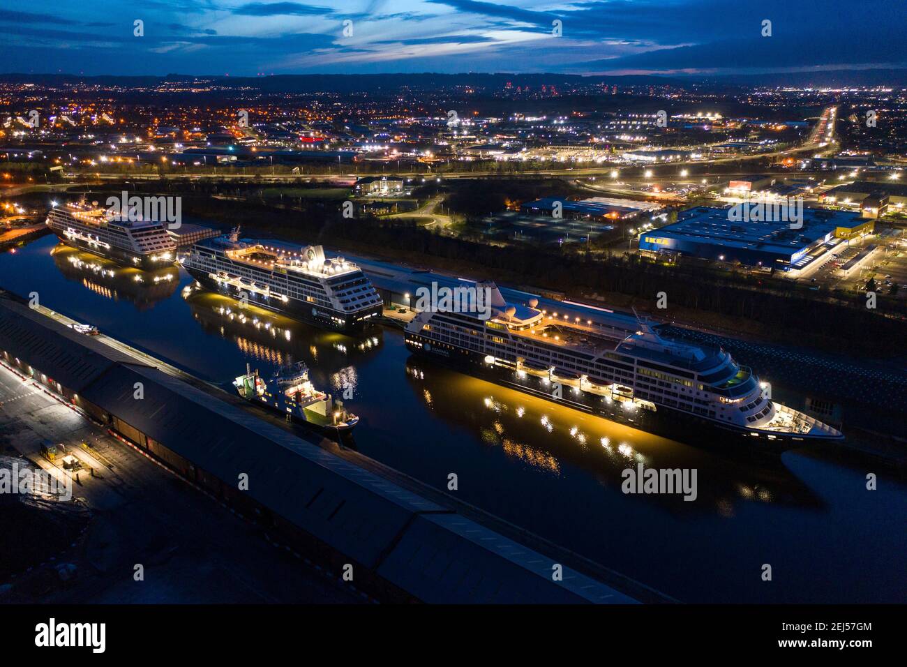 Glasgow, Scotland, UK. 21st Feb, 2021. Pictured: Aerial drone photography of the  three Amazara ruise ships (L-R) Amazara Pursuit; Amazara Quest; Amazara Journey, seen with their lights, have been berthed King George V Dock since the start of the coronavirus (COVID19) lockdown early last year. The evening skyline of Paisley and Glasgow Airport are visible with the M8 motorway. Credit: Colin Fisher/Alamy Live News Stock Photo