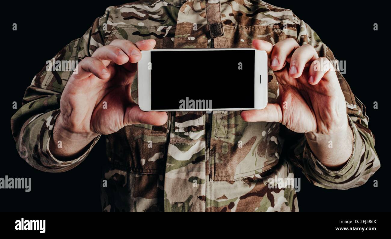 Photo of soldier in camouflaged uniform holding smartphone on black background. Stock Photo