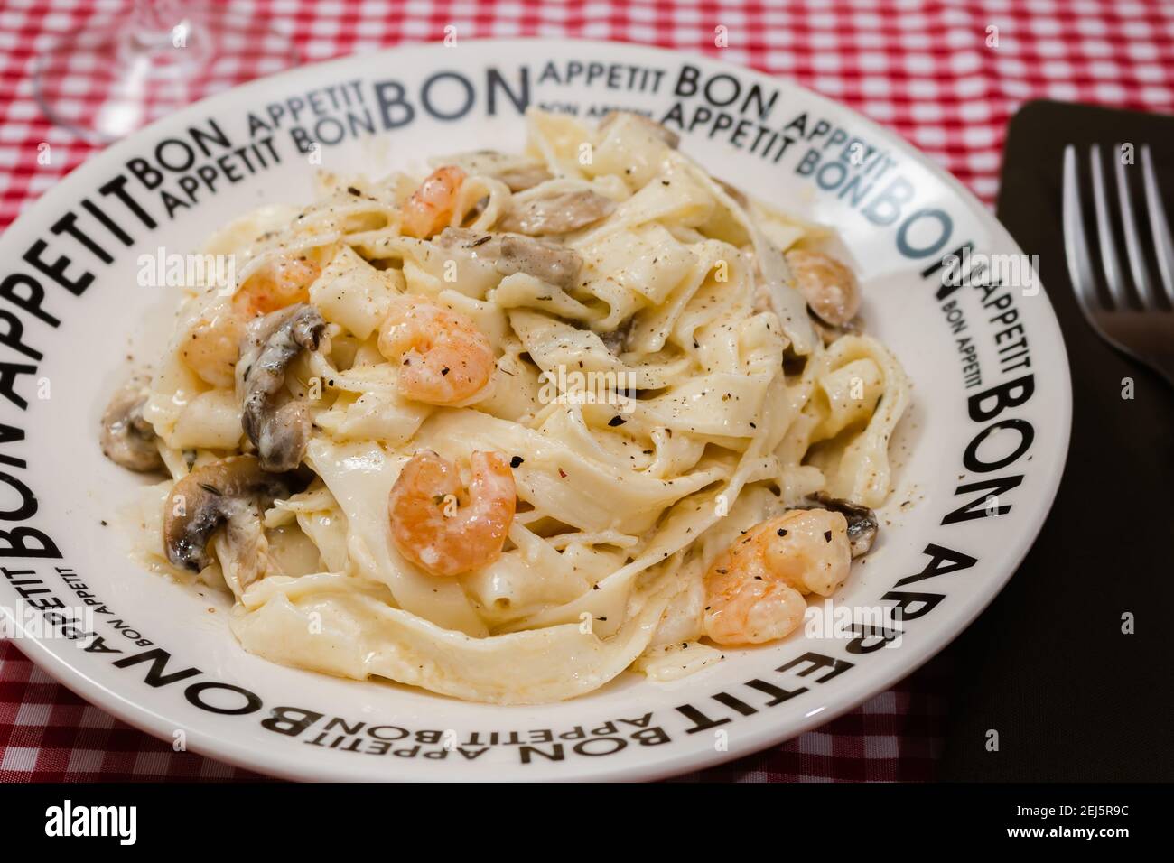 Delicious fettuccine with cream sauce of mushrooms and shrimp on a plate that says Bon Appetite with red checkered tablecloth. Italian cuisine. Stock Photo