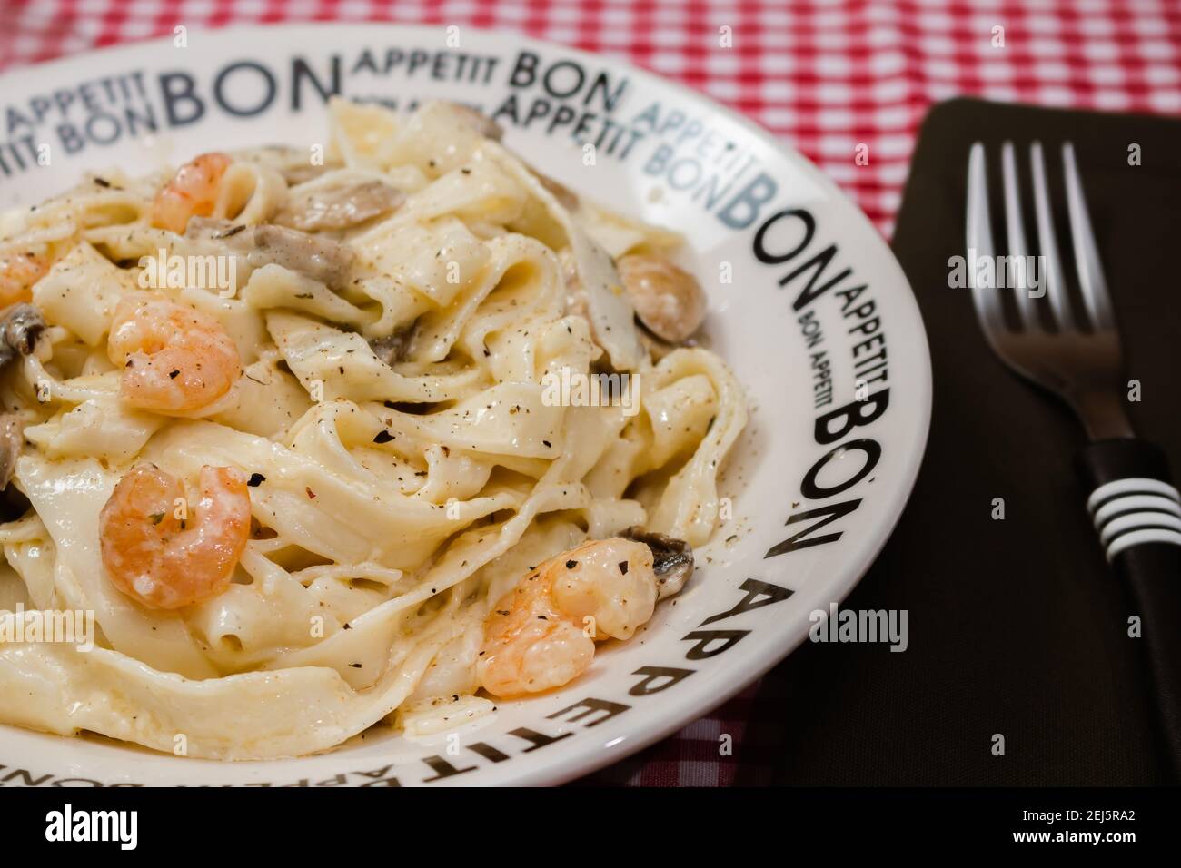 Delicious fettuccine with cream sauce of mushrooms and shrimp on a plate that says Bon Appetite with red checkered tablecloth. Italian cuisine. Stock Photo
