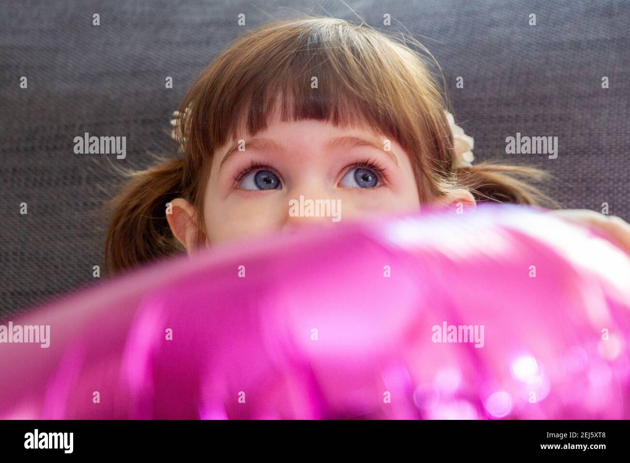 A cute, brown-haired, blue-eyed baby girl sitting on a sofe and hading her face behind a big pink balloon. Stock Photo