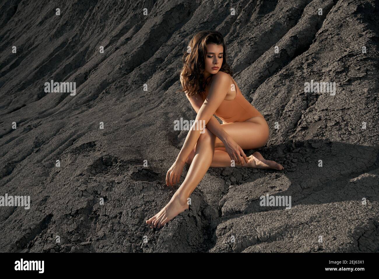 Side view of astonishing beautiful caucasian female model wearing beige body posing in dry empty quarry in hot summer sunny day. Young barefoot stylish woman sitting on black sand outdoors. Stock Photo