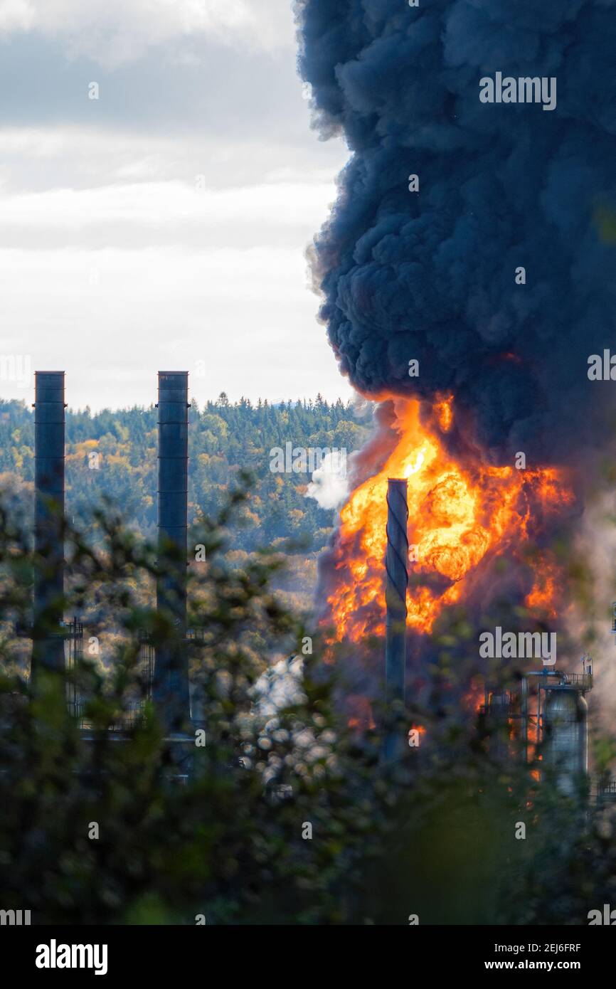 Large fire after an explosion at an oil refinery. Seen from a distance, one smoke stack silhouetted by bright flame. Thick black smoke rises. Stock Photo