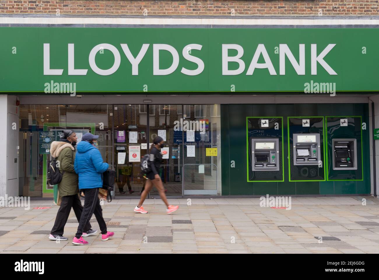 customers wearing face mask outside branch entrance to Lloyds Bank, White letter banner in green background and  two cash machines on the wall Stock Photo