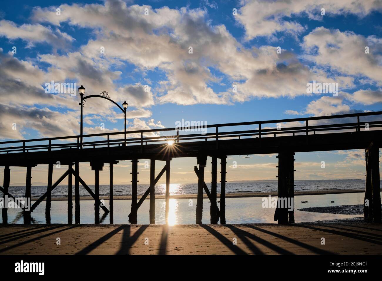 White Rock Pier is silhouetted against the sea and blue sky with light cloud.  Setting sun appears as star on pier Stock Photo