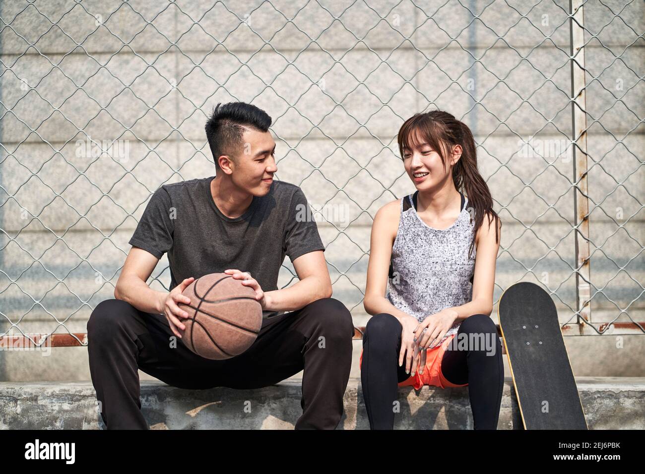 young asian adult male basketball player and female skateboarder sitting on court side talking chatting Stock Photo