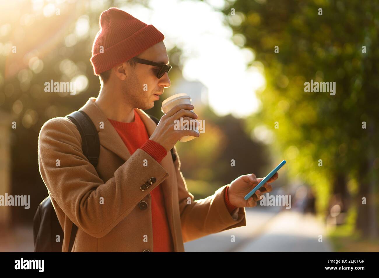 Smiling hipster man wear sunglasses using cellphone standing at the street drinking takeaway coffee  Stock Photo