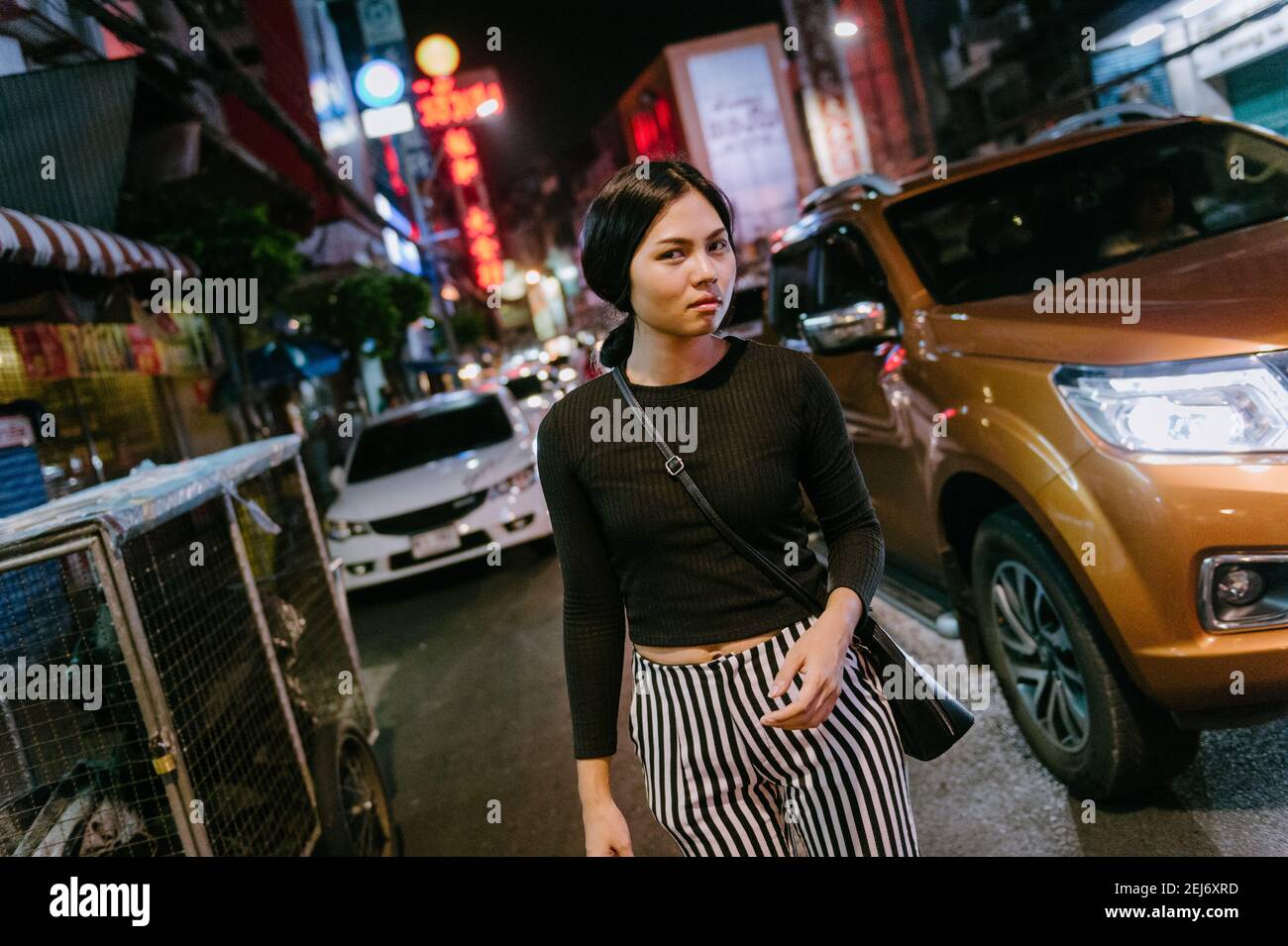 Young beautiful Asian girl in a busy street at night, walking while looking away from the camera. City lights and car in the background. Stock Photo