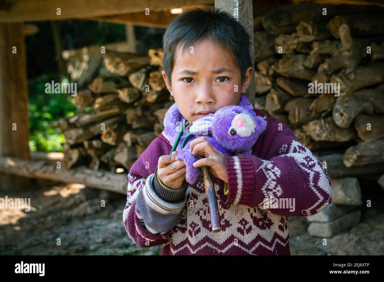 Closeup of a small boy holding his purple toy outside his house in the village with firewood in background. Stock Photo