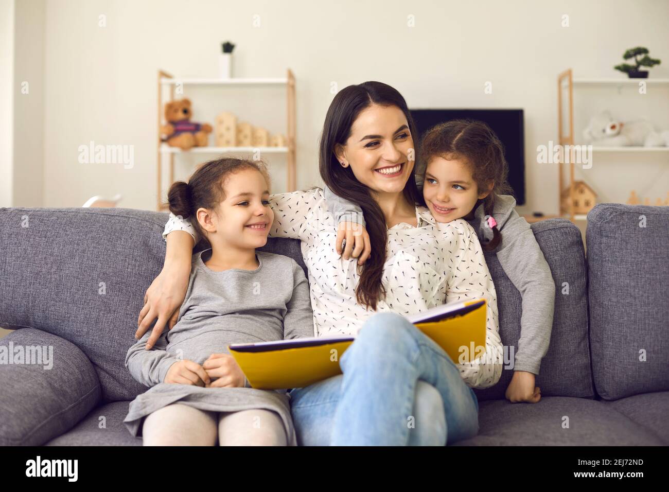 Smiling cheerful mother and small daughters sitting on sofa with book, hugging and looking aside Stock Photo