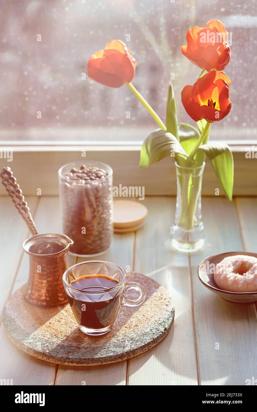 Oriental coffee in traditional Turkish copper coffee pot with flowers on window sill. Wooden windowsill with orange tulips and hyacinth flower pot Stock Photo