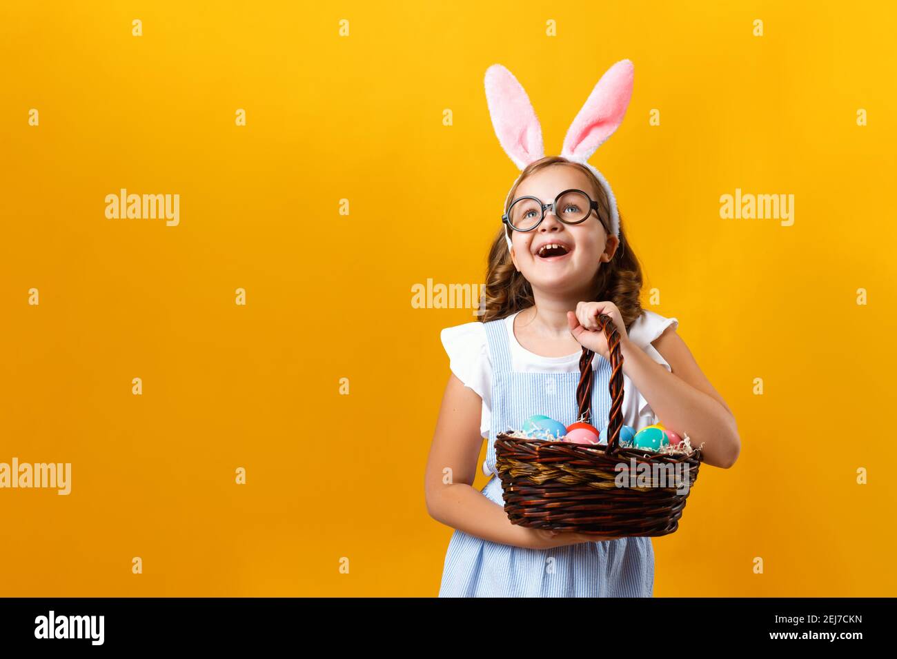 Cute little girl holding an Easter basket with eggs on a yellow background. Happy kid lifted his head and looks up. Copy space. Stock Photo