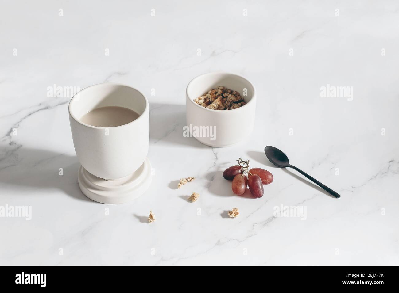 Breakfast, snack still life scene. Cup of coffee, red grapes fruit on white marble table in sunlight. Bowl with muesli, granola and oat cereals and Stock Photo