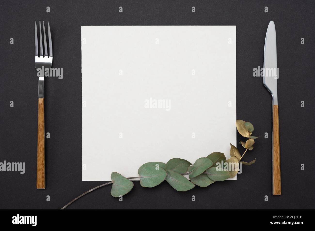 Mockup menu, recipe or cookbook. knife, fork, square paper mockup decorated with gold eucalyptus branch on a black table. Stock Photo