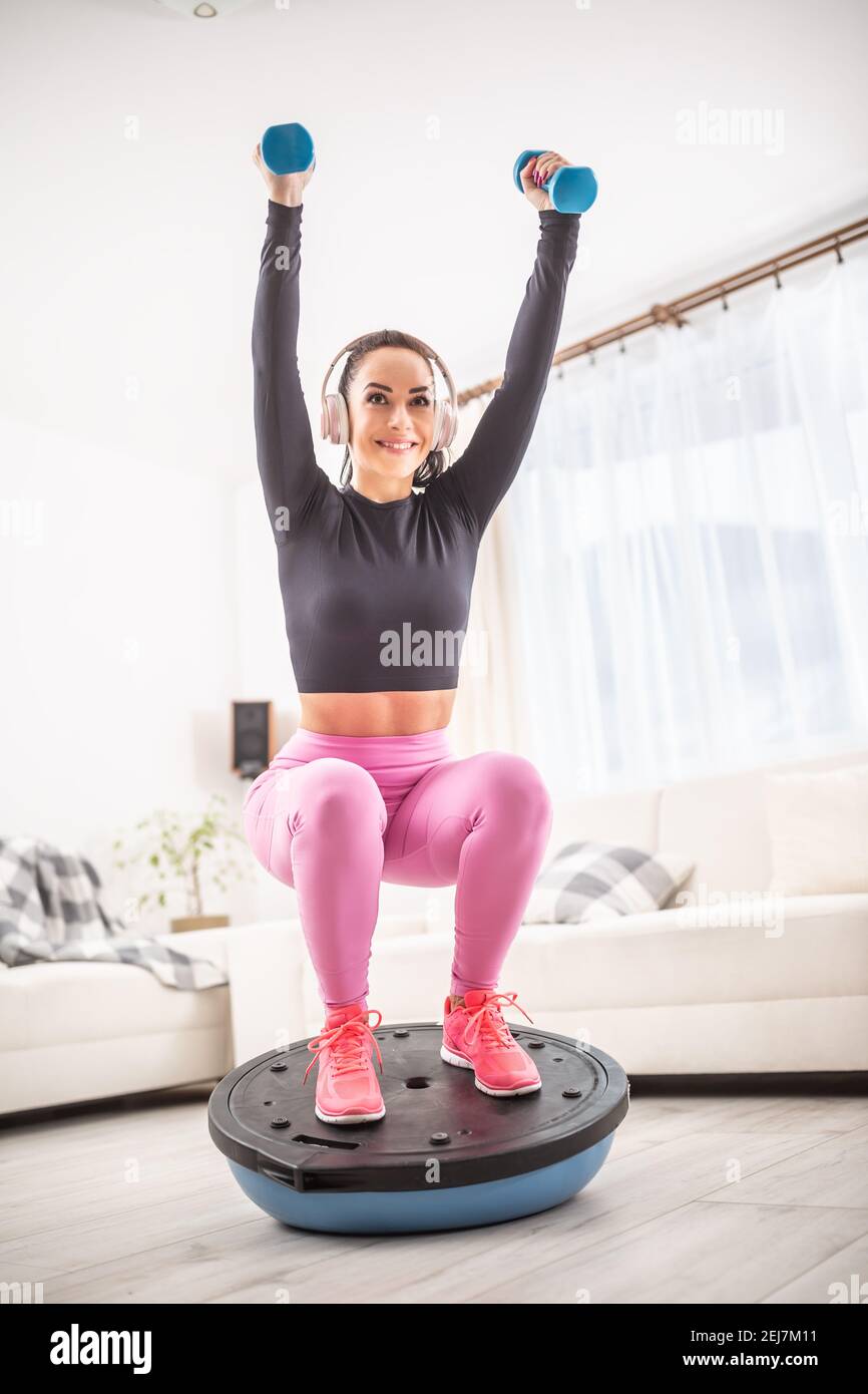 Fit beautiful woman in a training outfit and headphones squats at home on a balance ball holding dumbells upwards in stretched arms. Stock Photo