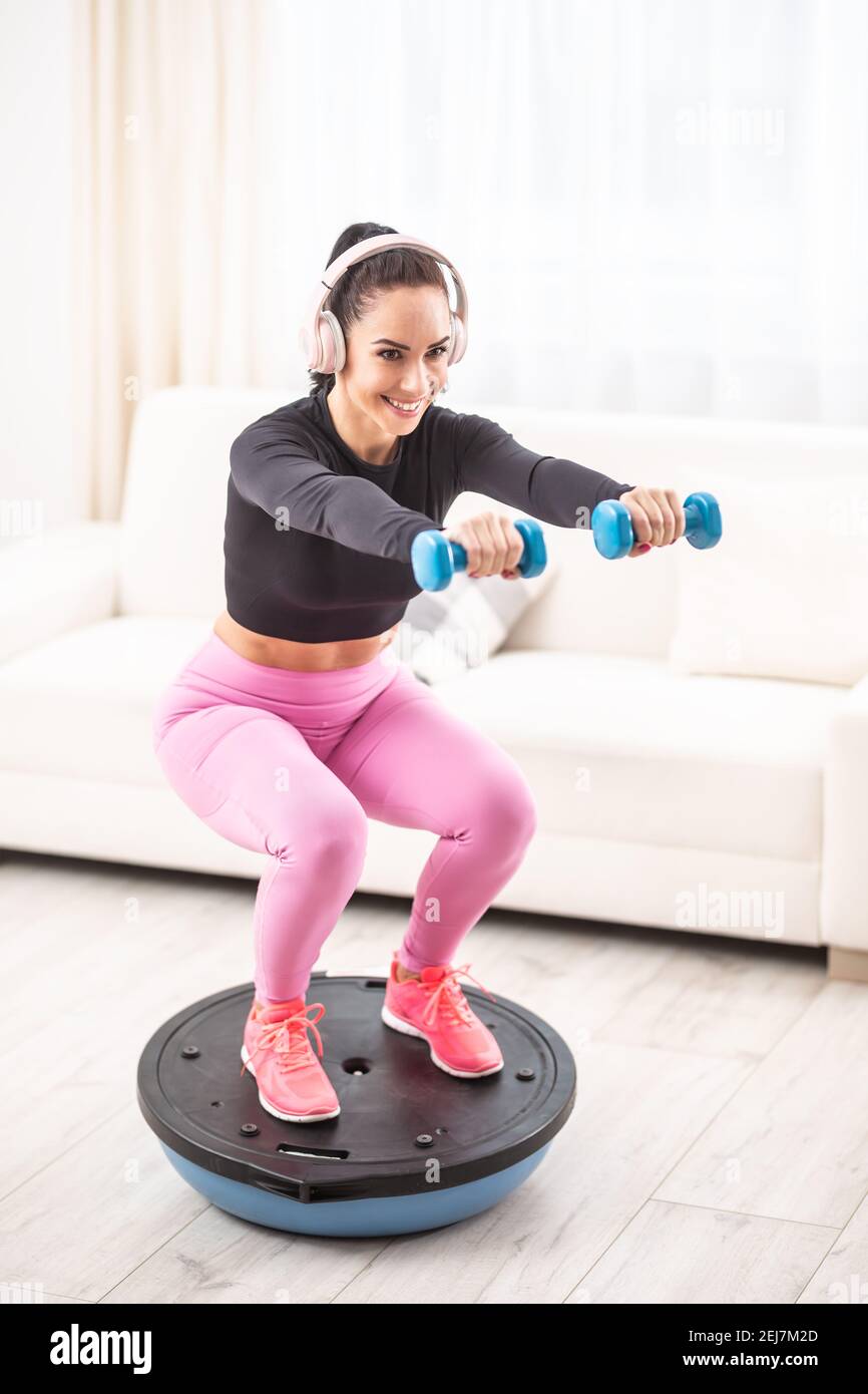 Fit beautiful woman in a training outfit and headphones squats at home on a balance ball holding dumbells. Stock Photo