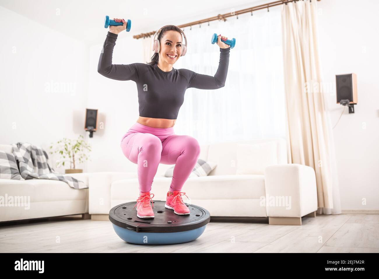 Fit beautiful woman in a training outfit and headphones squats at home on a balance ball holding dumbells upwards. Stock Photo