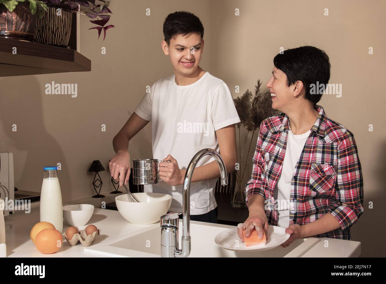 Happy smiling mother and son on kitchen. Teenage boy helping to cook food, mom washing the dishes. Relationship between parents and children Stock Photo