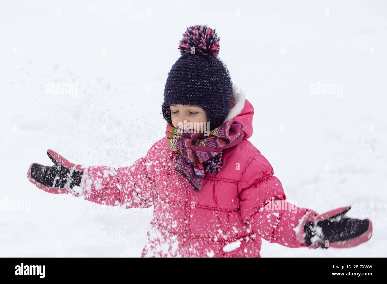 A cute little girl in a pink jacket and a hat is playing in the snow ...