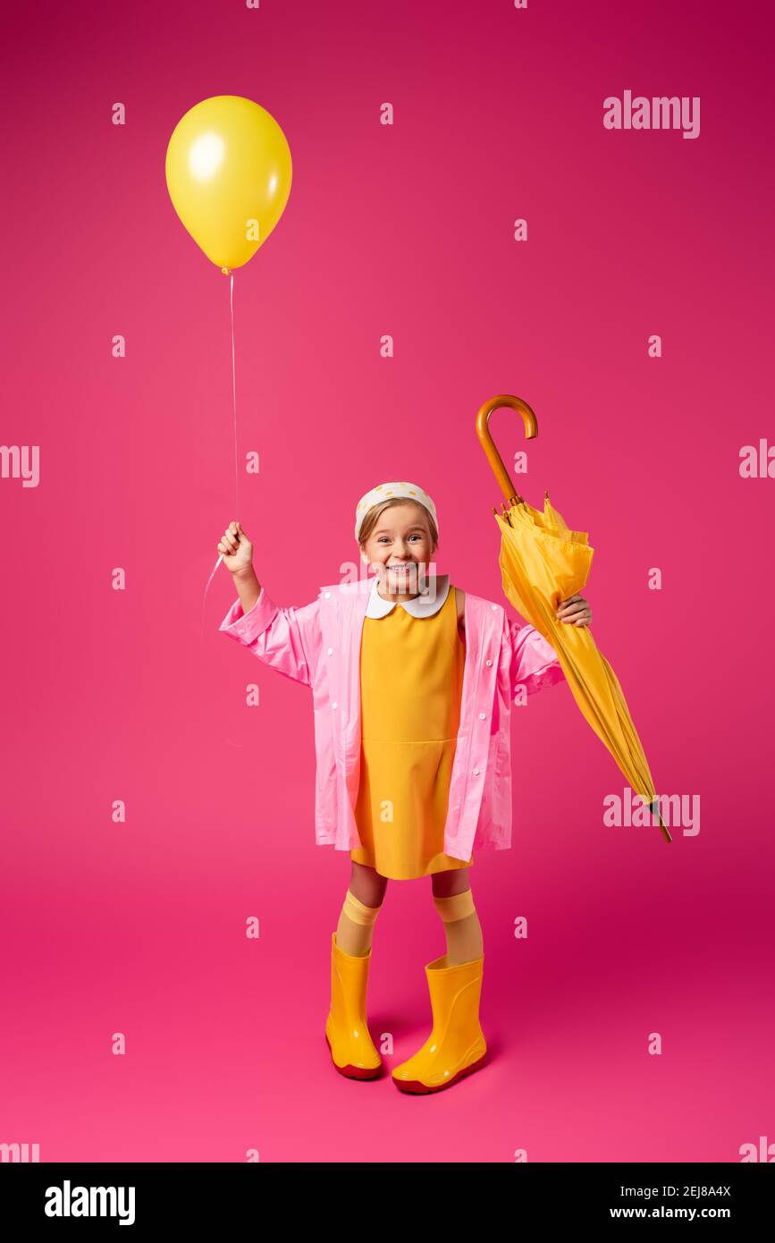 full length of happy girl in raincoat and rain boots holding balloon and yellow umbrella on crimson Stock Photo