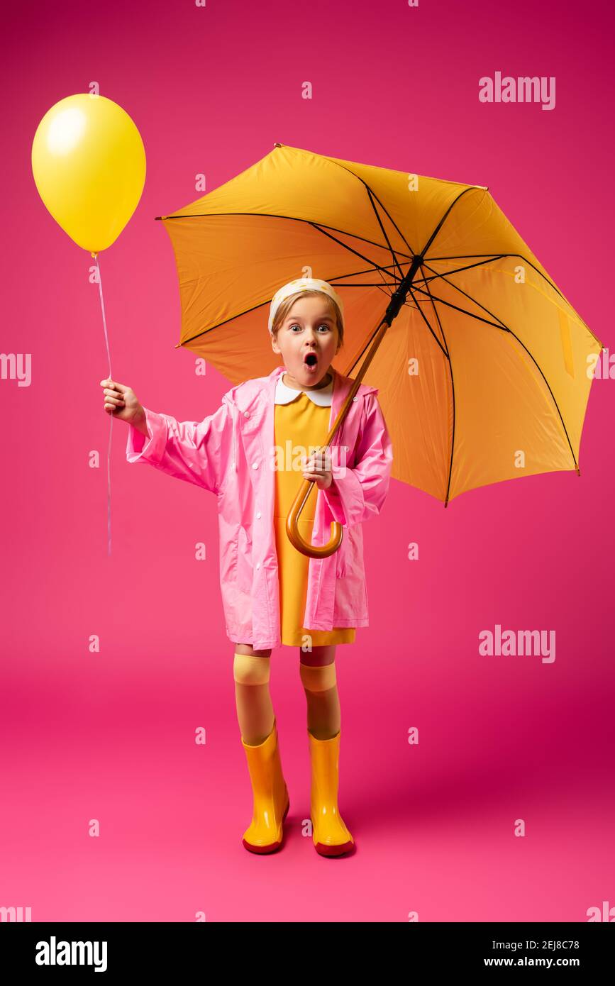 full length of amazed child in raincoat and rubber boots holding balloon and yellow umbrella on crimson Stock Photo