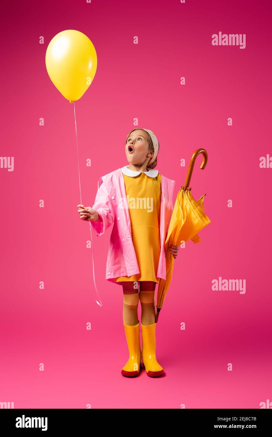 full length of amazed girl in raincoat and rubber boots holding balloon and yellow umbrella on crimson Stock Photo