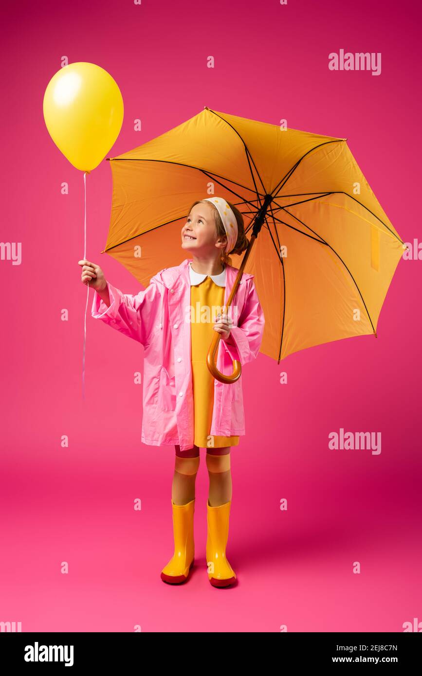 full length of cheerful girl in raincoat holding balloon and yellow umbrella on crimson Stock Photo