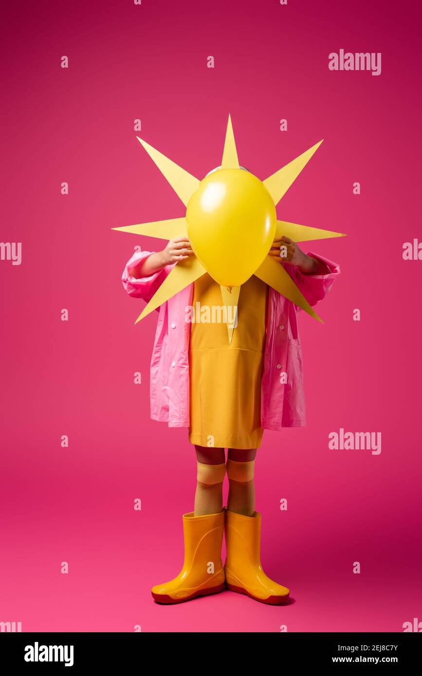full length of girl in raincoat and rubber boots covering face while holding decorative sun with balloon on crimson Stock Photo