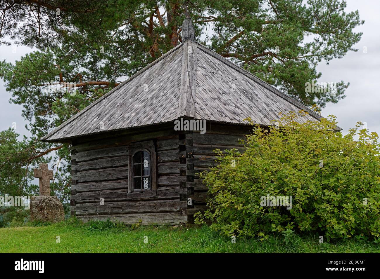 View of the wooden chapel of the Archangel Michael with a stone cross at the entrance to the Savkino settlement in Pushkinskiye Gory. Stock Photo