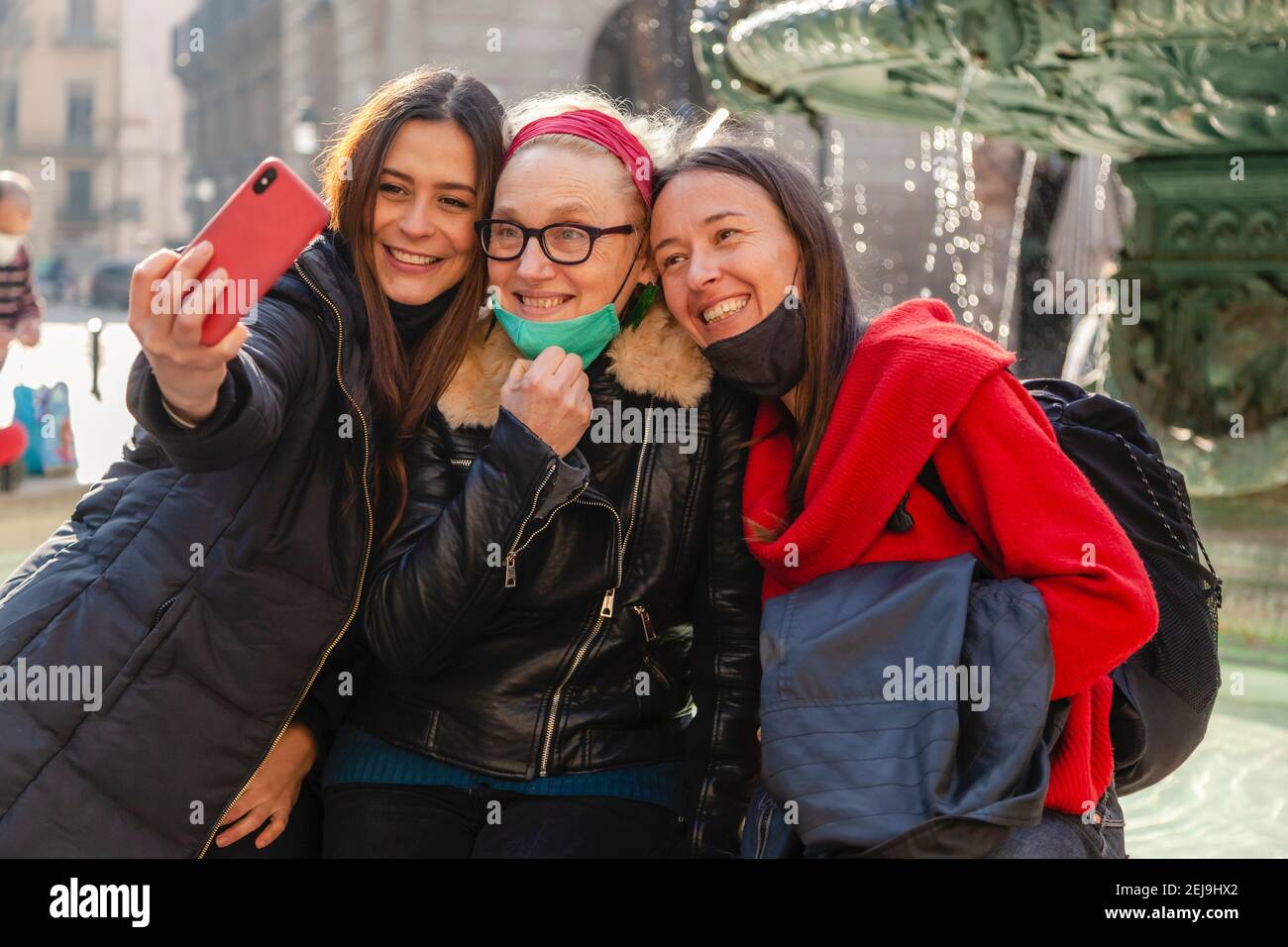 Three women friends capturing a selfie with a smart phone, smiling and sitting close together in urban environment Stock Photo