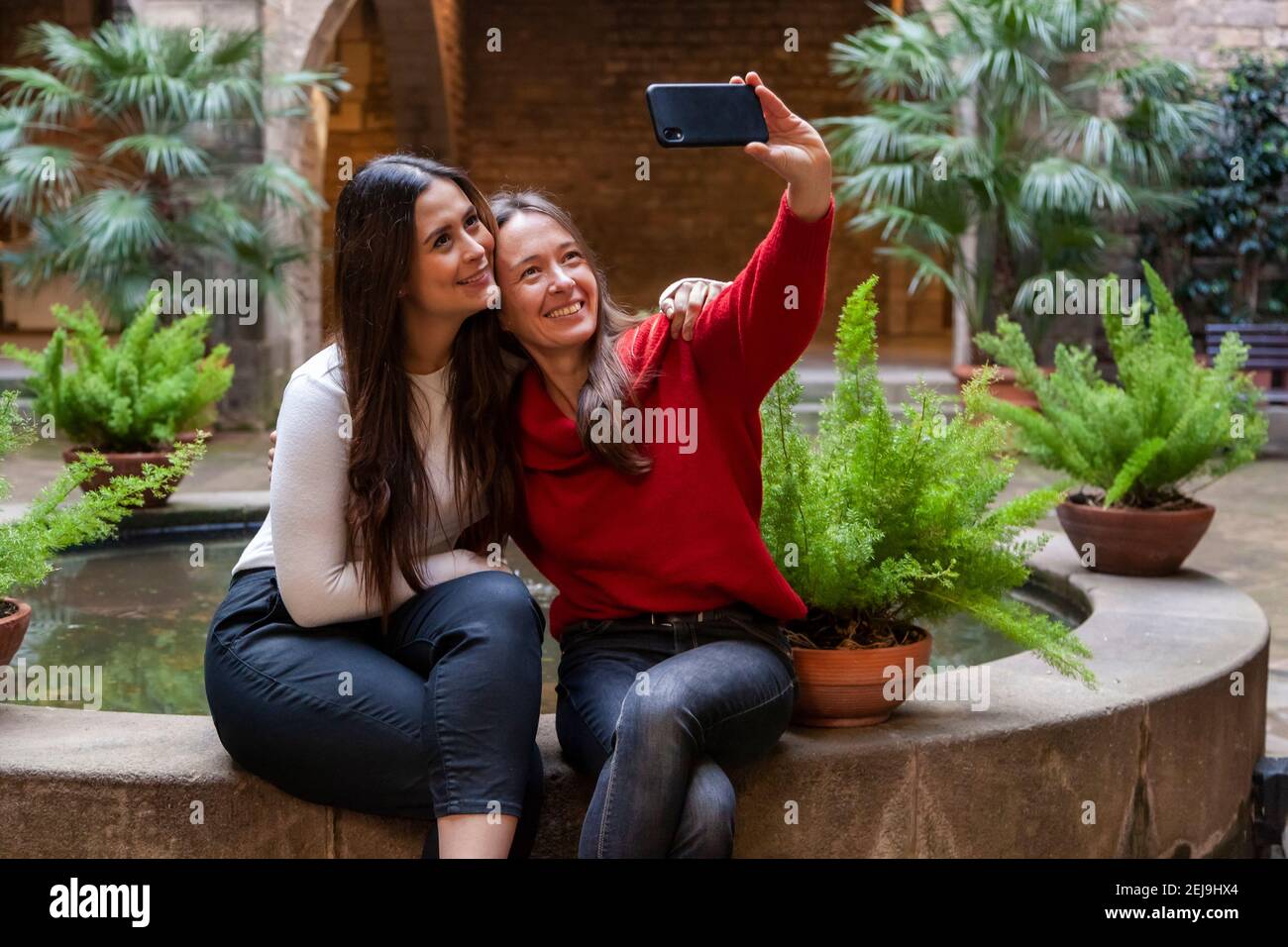 Two women friends capturing a selfie with a smart phone, smiling and sitting close together in urban environment Stock Photo