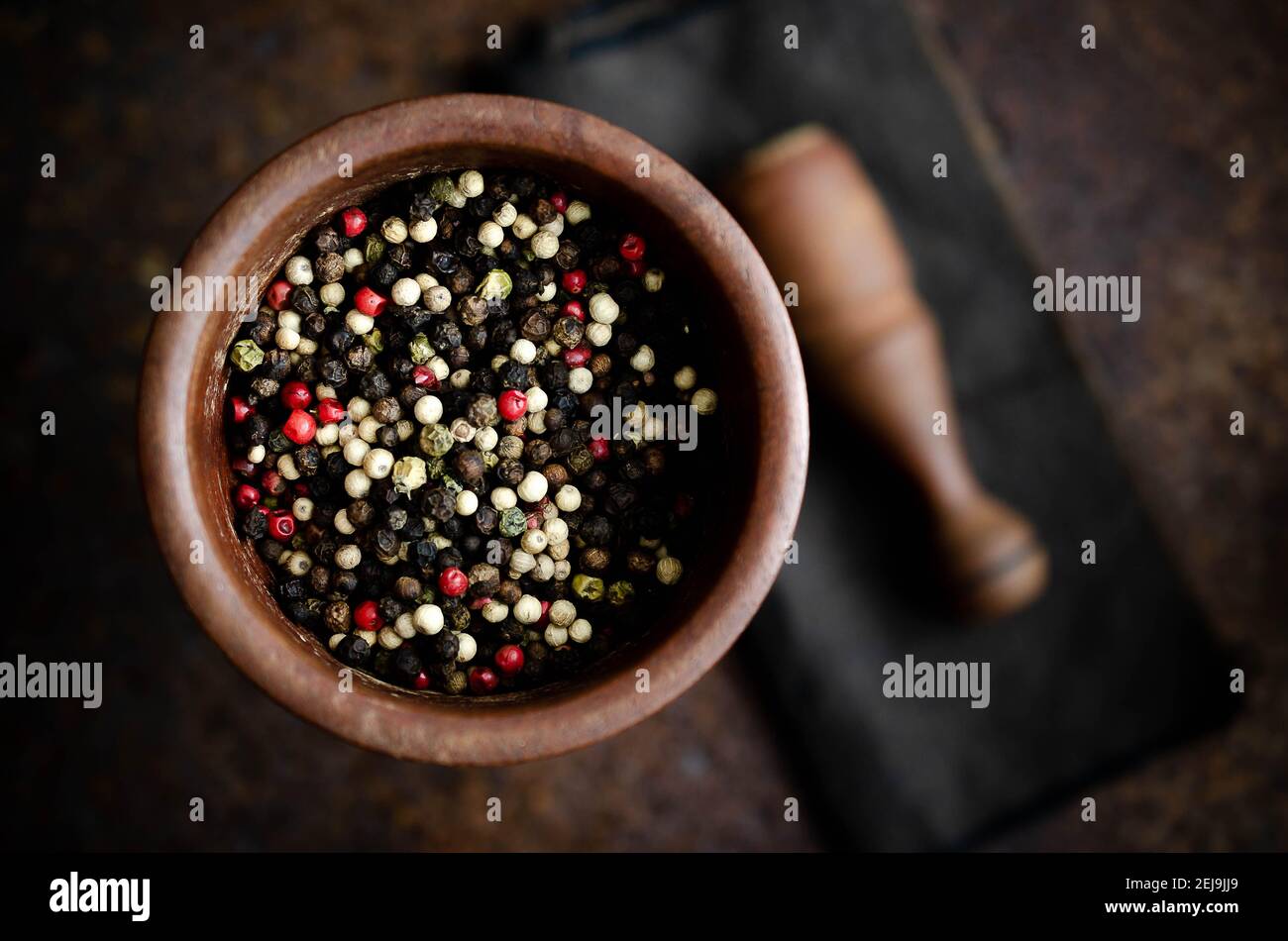 Black, white, green, and pink peppercorns in a wooden mortar with pestle on a brown napkin and dark backdrop. Stock Photo