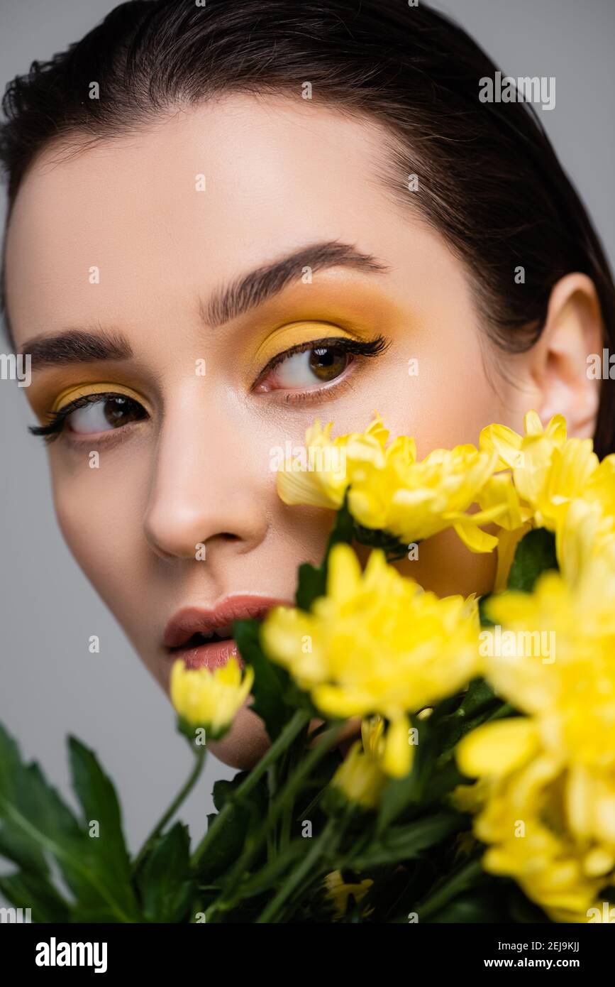 young brunette woman with makeup near blooming yellow flowers on blurred foreground isolated on grey Stock Photo