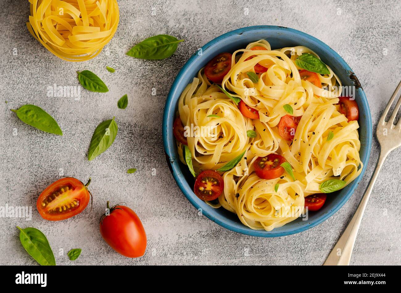 Fettuccine with cherry tomatoes and basil leaves in a blue plate on a grey backdrop with one and a half tomato, basil leaves and a nest of raw pasta. Stock Photo