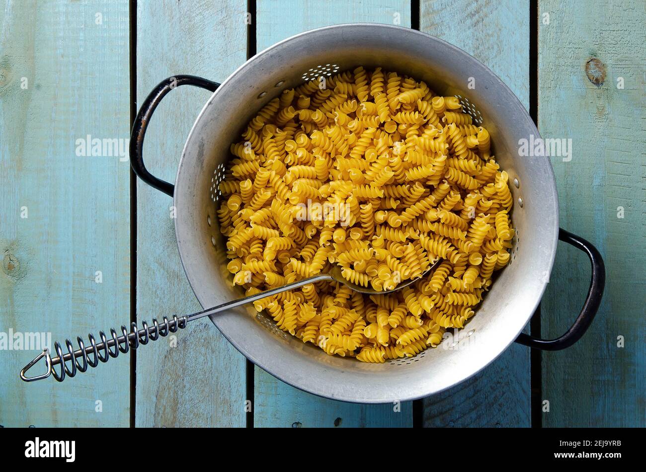 Uncooked spiral pasta in a strainer with metal spoon, on a light blue backdrop. Top view. Stock Photo