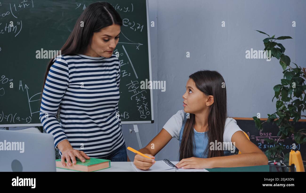 mother standing near upset daughter writing on notepad while doing homework Stock Photo