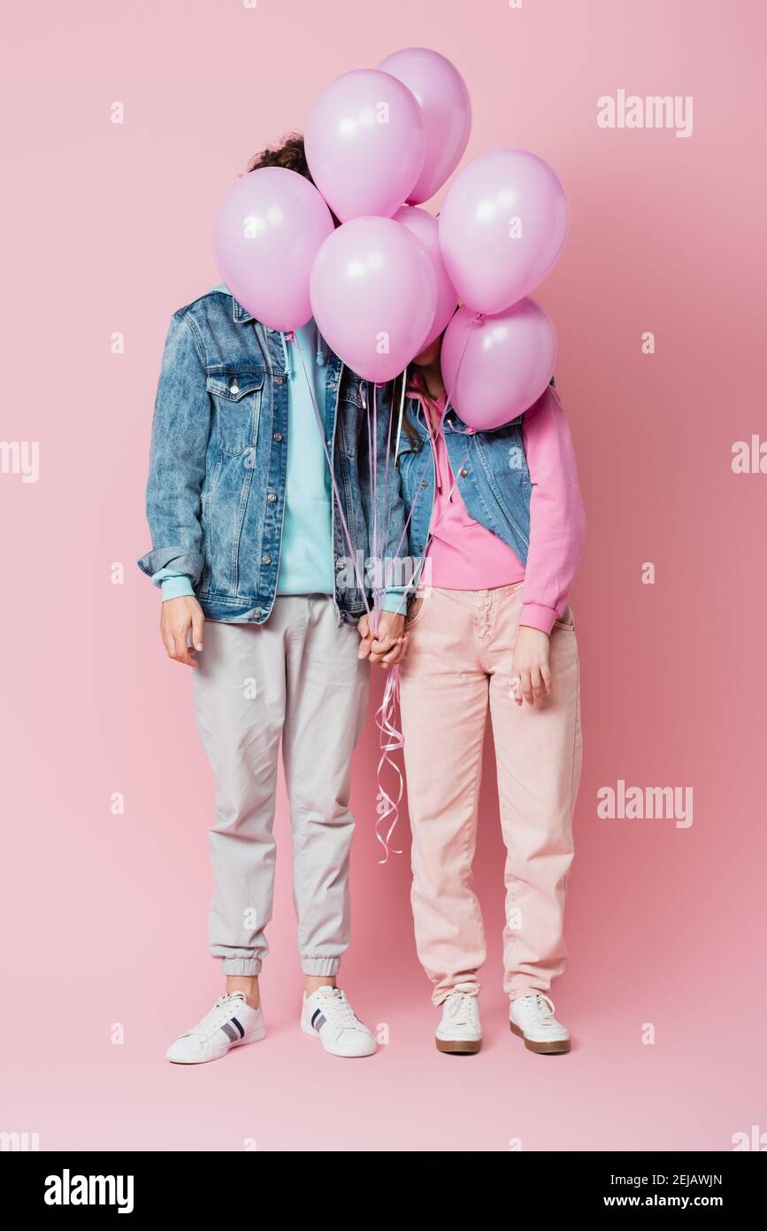 Teen couple holding hands while balloons covering faces on pink background Stock Photo