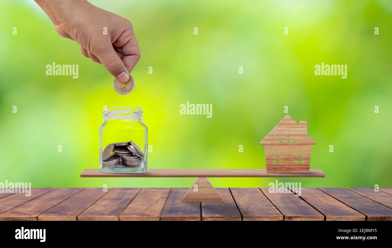 Hands that are putting coins into money saving bottles and model wooden houses on wooden scales in savings ideas for buying a new home or real estate. Stock Photo