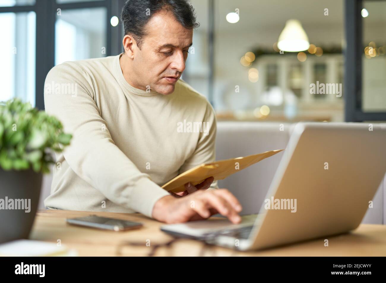 Busy latin middle aged businessman holding document envelope while working using laptop at home. Telework, business concept Stock Photo