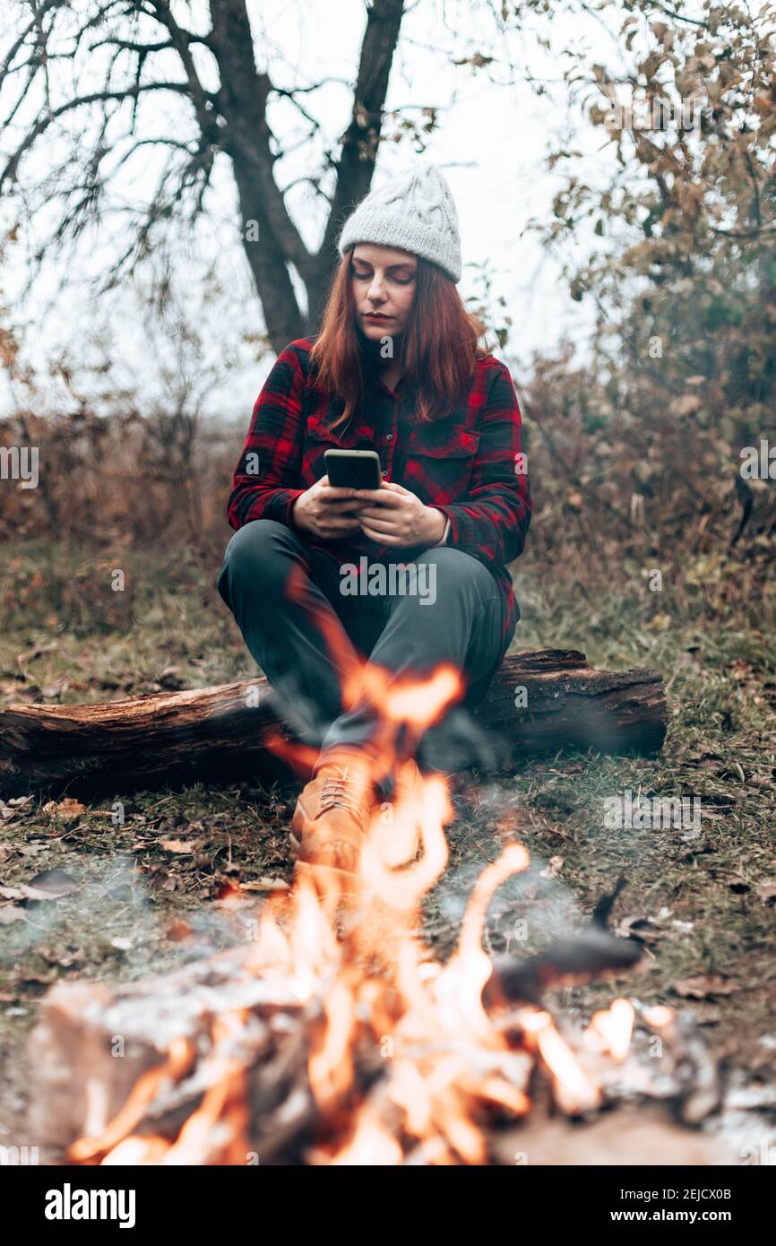 A young traveler girl in a plaid shirt and hat uses a smartphone sitting by the fire near the camp. Woman enjoying relaxing in the forest Stock Photo