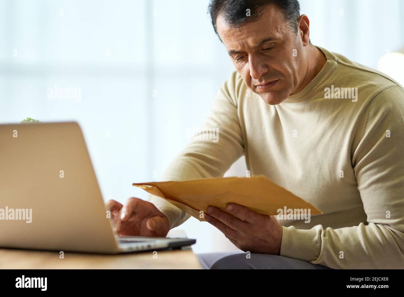 Portrait of busy latin middle aged businessman looking at the envelope, using laptop while working from home. Telework, business concept Stock Photo