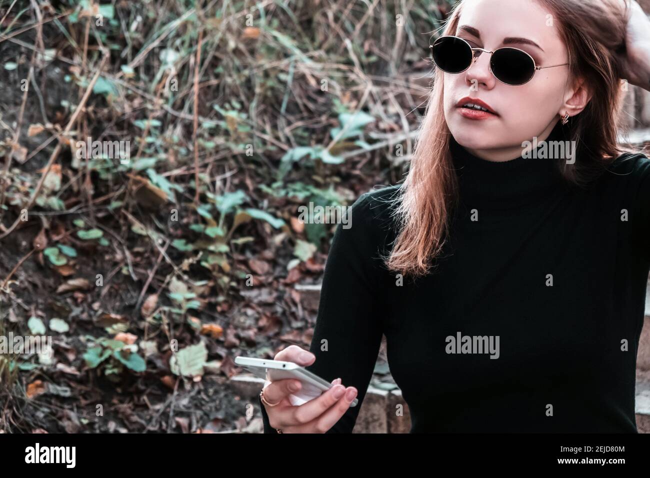 Portrait of young stylish modern woman in sunglasses and black turtleneck sitting on the steps in the park holding mobile phone in her hand. Woman enj Stock Photo