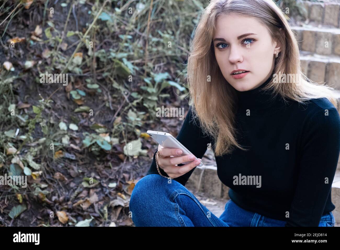 Portrait of a young attractive serious blue eyed woman in a black turtleneck looking into the camera sitting on the steps outside holding a mobile pho Stock Photo