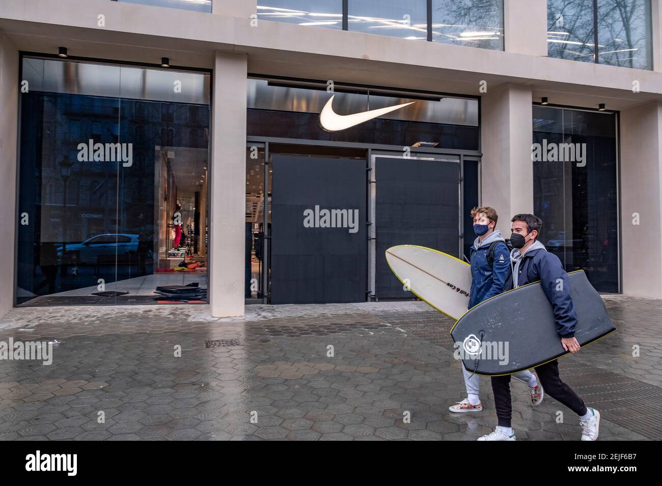 Barcelona, Spain. 22nd Feb, 2021. Two young men with surfboards walk past Nike  store in Passeig de Gràcia, seen with anti-vandalism protections in their  windows.More than 50 stores have suffered damage to