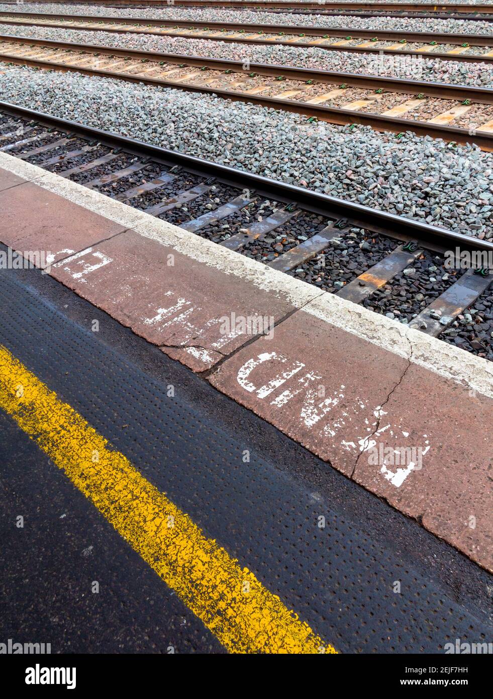 Yellow warning stripe and faded Mind The Step notice on UK railway station platform with railway tracks in background. Stock Photo