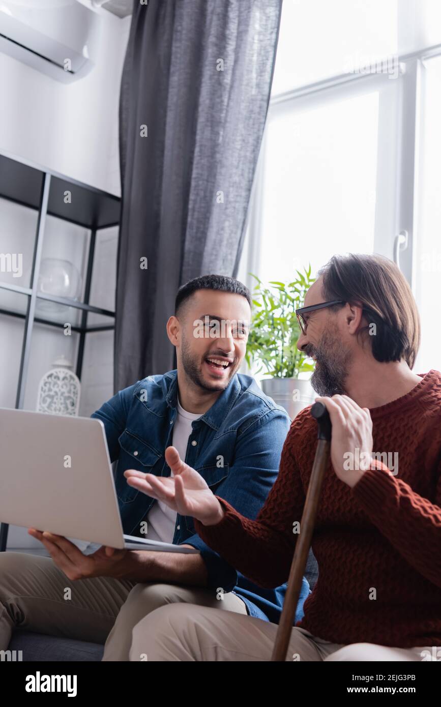 bearded man with walking stick pointing at laptop near excited hispanic son Stock Photo