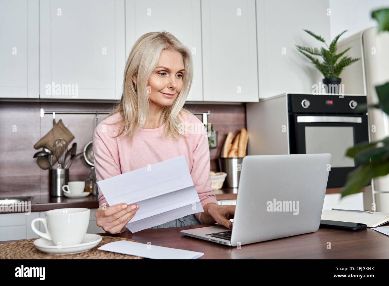 Middle aged mature woman holding paper bill or letter using laptop at home. Stock Photo