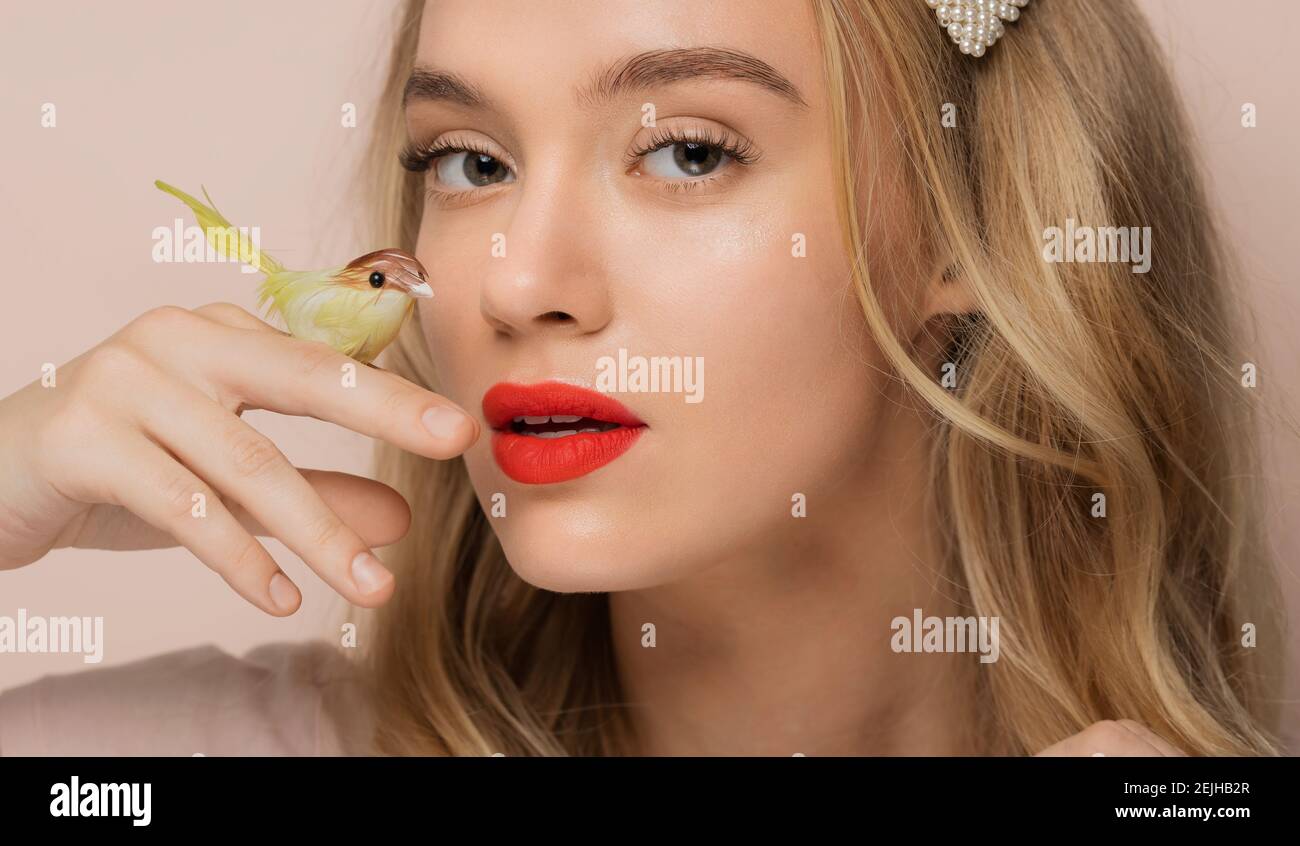 Young beautiful blonde girl holding artificial bird on finger on pink background. Eyelash extension beauty procedure concept without animal tests. Stock Photo