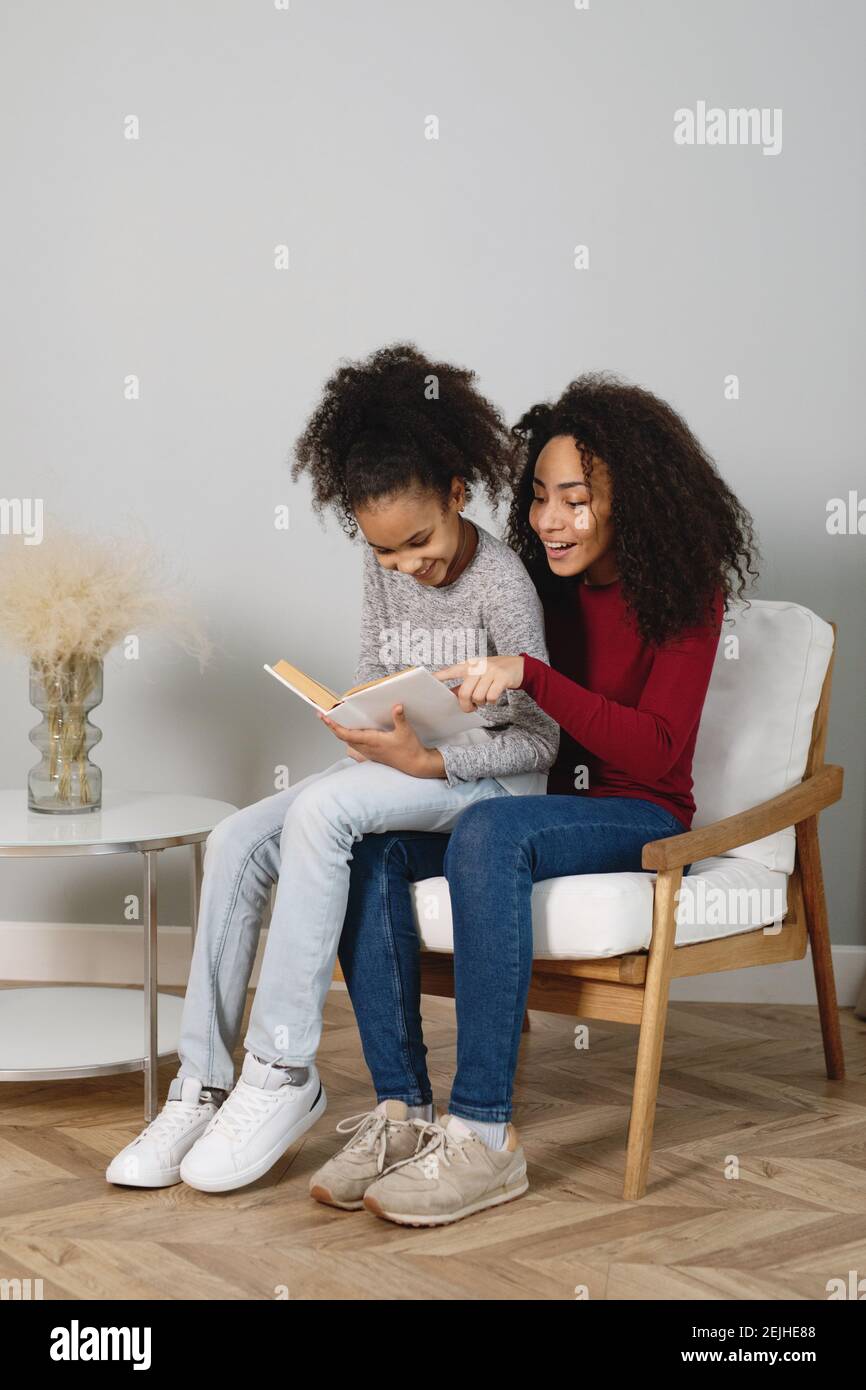 African American mother with her cute daughter reading a book sitting in a chair. Stock Photo